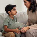 Boy holds a cushion on a sofa and tentatively questions his parent, who listens calmly at his eye level.